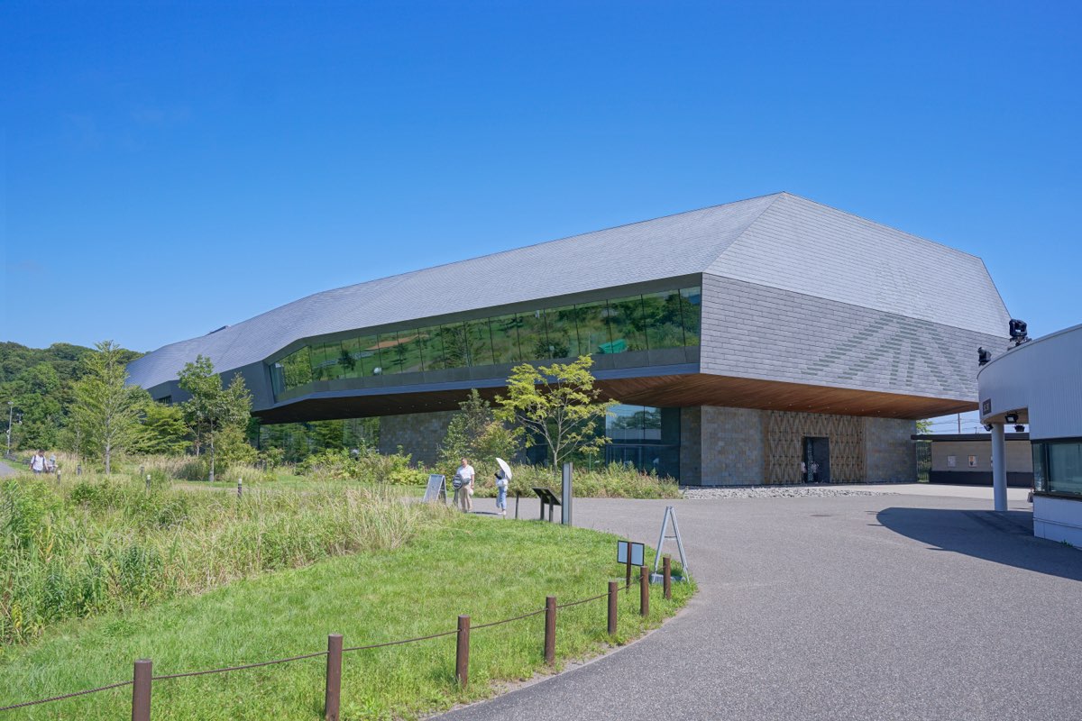 The National Ainu Museum at Upopoy in Shiraoi, Hokkaido, a long modern building with a grey roof, seen from a paved path beside grass under a clear summer sky in 2026