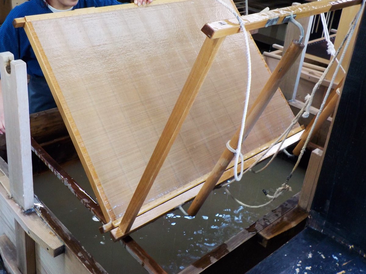 A large bamboo papermaking screen in a wooden frame, hung from cords, being tilted up out of a vat of water