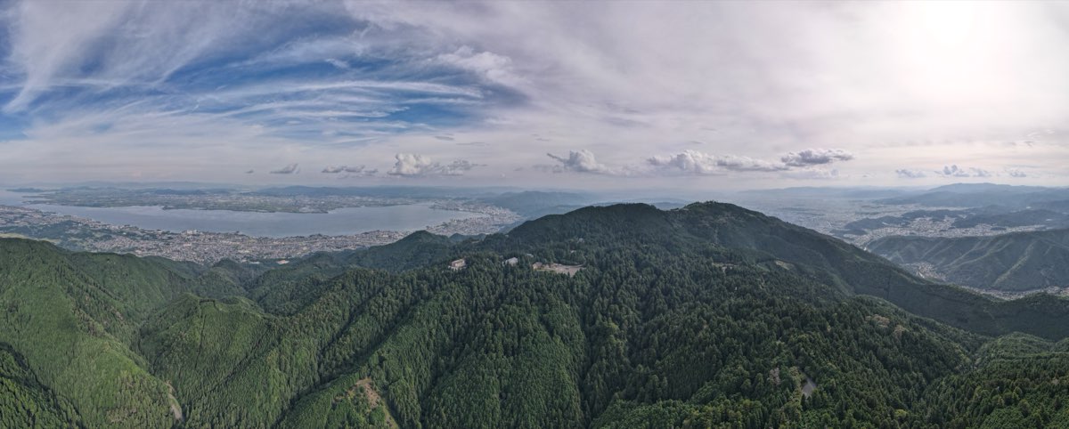 木々に覆われた比叡山の尾根を空から見渡した横長の写真。木々の間に建物がいくつか見え、左手には琵琶湖とその岸に広がる大津の街並み、上には雲の多い空が広がる