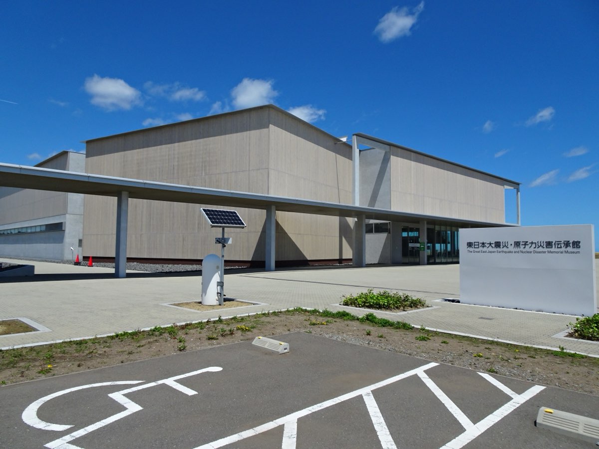The pale-grey building of the Great East Japan Earthquake and Nuclear Disaster Memorial Museum in Futaba, with a long canopy over the entrance, a white sign bearing the museum's name in Japanese and English, and a paved forecourt and car park under a blue sky