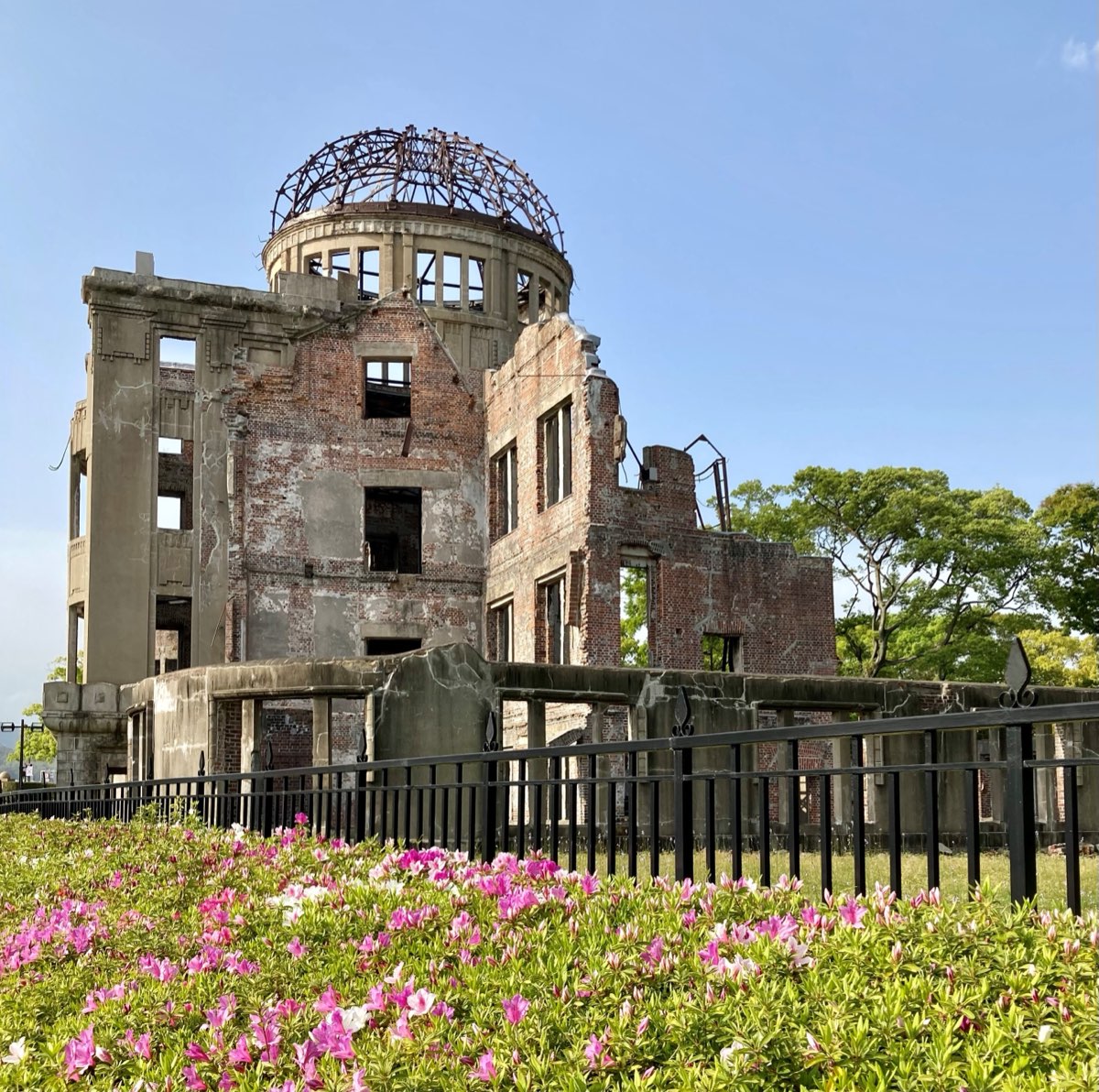 青空の下の原爆ドーム。窓の抜けたれんがとコンクリートの壁と、骨組みだけになったドームが、黒い鉄の柵の向こうに見える。手前にはピンクのツツジが咲いている