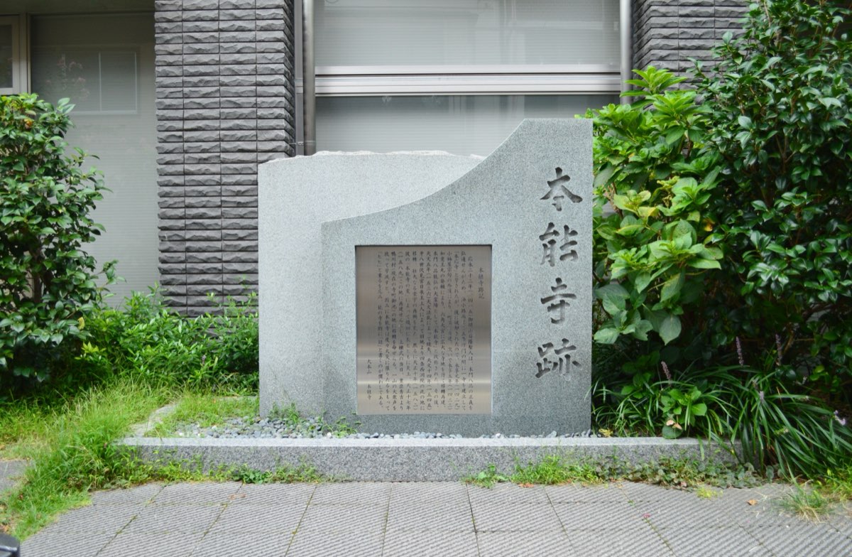 現代の建物の前、植え込みに囲まれて立つ灰色の御影石の石碑。右側に「本能寺跡」の文字が縦に彫られ、中央に文章を刻んだ金属の銘板がはめ込まれている