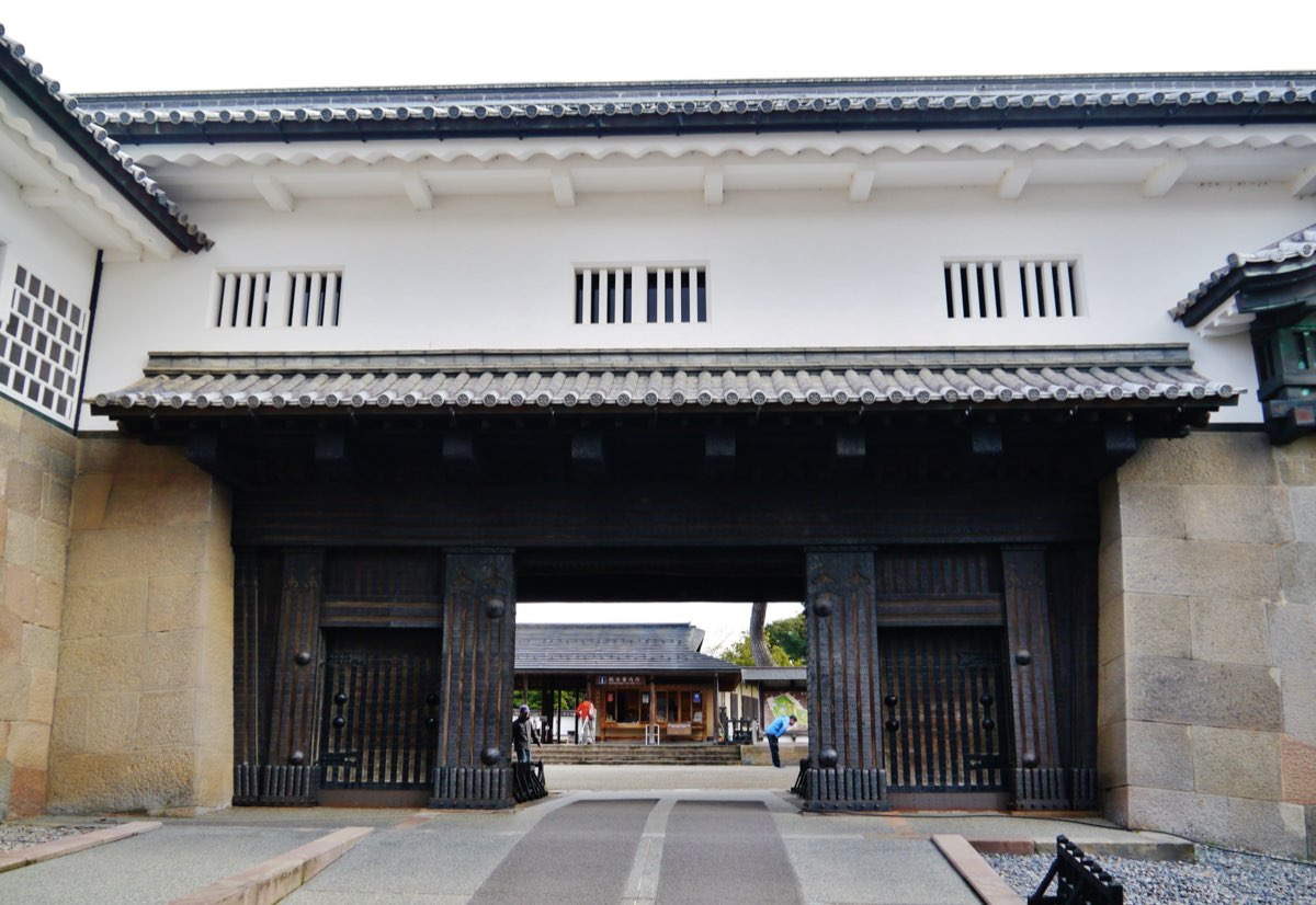 正面から見た金沢城の石川門。切り石を積んだ石垣の間に黒っぽい木の大きな門があり、その上に格子窓のある白壁の建物と灰色の瓦屋根が載る。開いた門の奥へ舗装された道が続き、その先の広場に小さな建物が見える