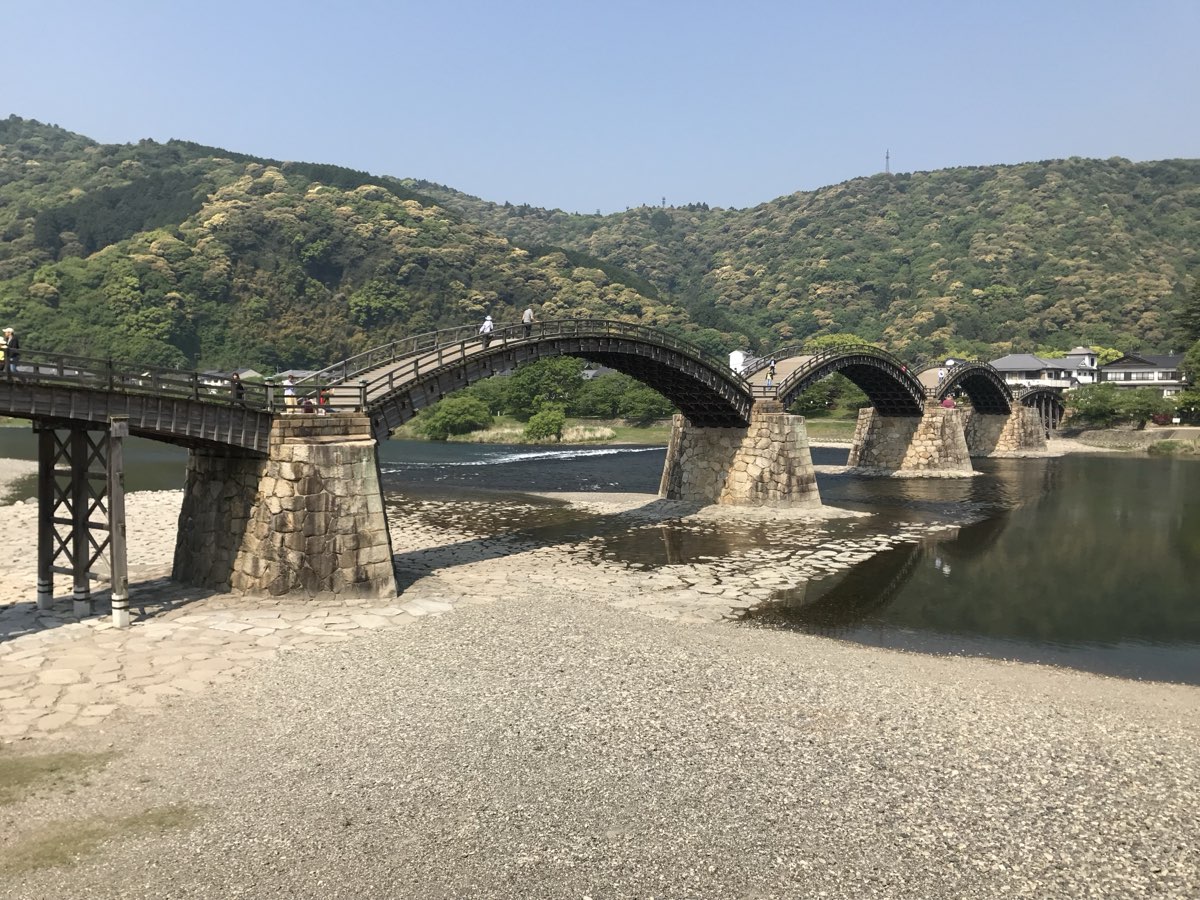 The Kintai Bridge seen from the dry riverbed, with a flat wooden span on wooden posts in front, then three high wooden arches resting on tall stone-faced piers, stone paving on the riverbed, and green hills behind under a clear sky