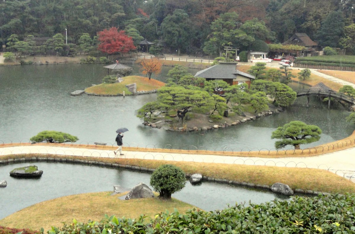 雨の日に高い所から見た後楽園の池。松の茂る島に小さな建物が建ち、もう一つの芝の島にはオレンジ色に色づいた木と傘のような形のあずまやがあり、対岸には紅葉した木が見える。右には反り橋、手前には二つの池の間の園路を傘をさして歩く人が一人、奥には木々の茂る丘が見える