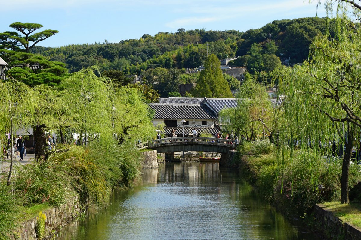 A narrow canal with stone banks running between green willows toward an arched stone bridge, with a white-walled building with dark tiled roofs behind the bridge, small figures of people on the bridge and the banks, and wooded hills under a blue sky