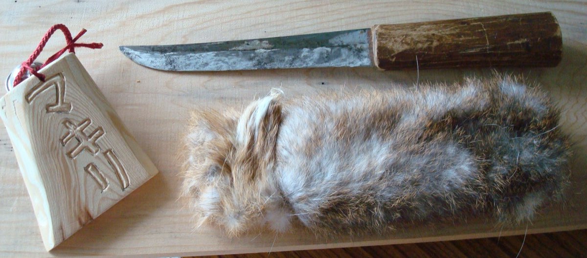 A worn knife with a wooden handle lying on a board beside a fur bundle and a wooden tag carved with the word makiri
