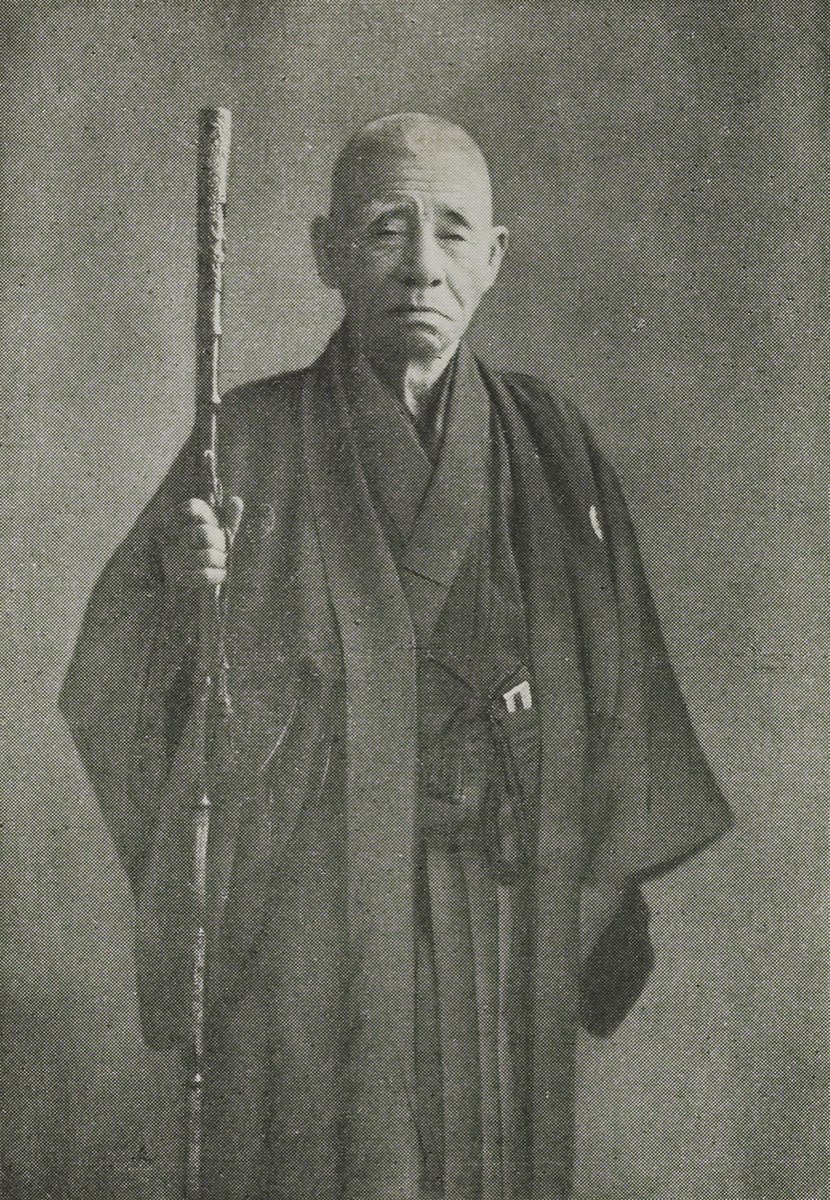 Black-and-white photograph of an elderly man with a shaved head standing in dark Japanese clothing with a family crest, holding a tall, knotted walking stick in his right hand, against a plain studio background