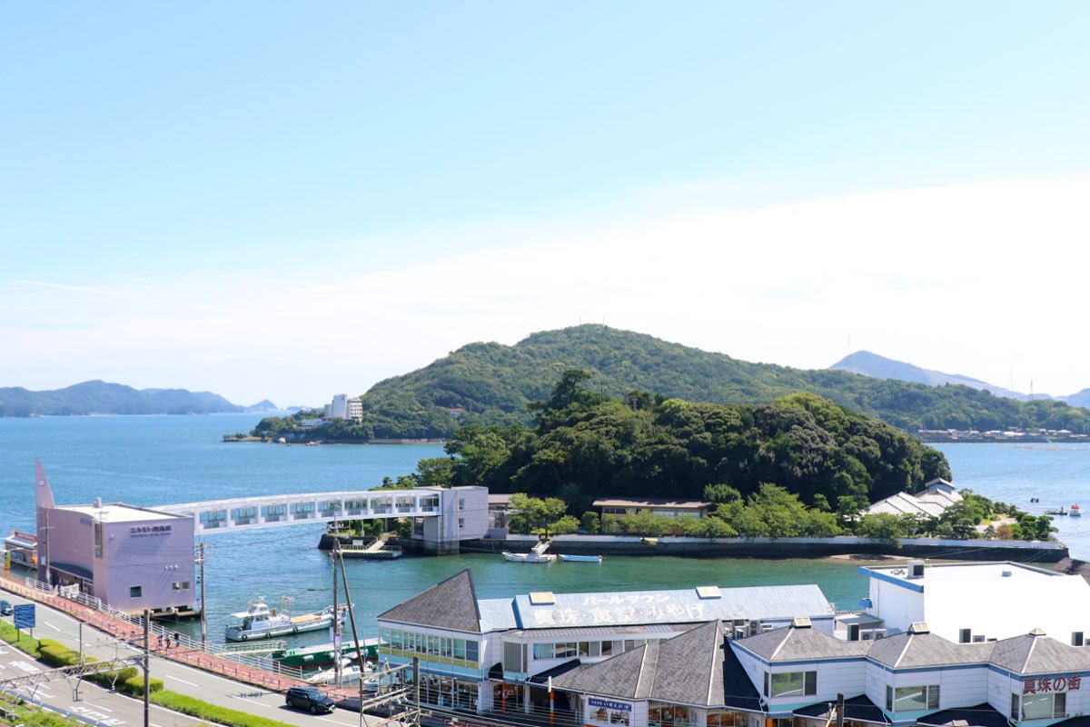 View over rooftops toward a small, tree-covered island in a calm blue bay, joined to the shore by a covered footbridge, with buildings at its water's edge, green hills behind and a clear sky above