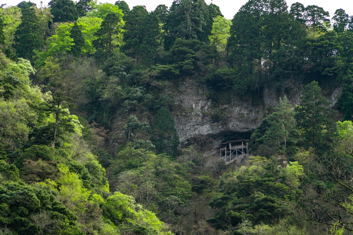 A forested mountainside in fresh spring green with a grey rock cliff in the middle; in a dark hollow at the foot of the cliff stands a small wooden hall raised on thin pillars