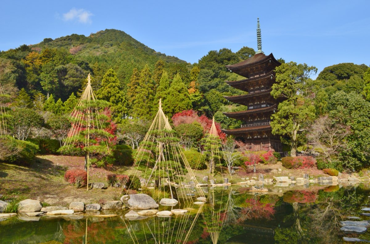 The dark wooden five-storied pagoda of Rurikōji rising among trees on the right, with a pond and garden in front, pine trees supported by rope frames, red autumn leaves and a wooded hill under a blue sky