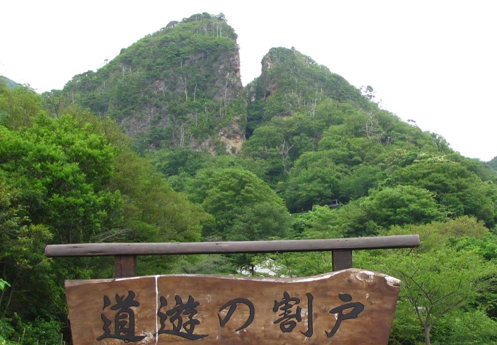 A forested mountain ridge split by a deep V-shaped cleft into two rocky peaks, seen above green trees, with a wooden sign reading 道遊の割戸 in the foreground