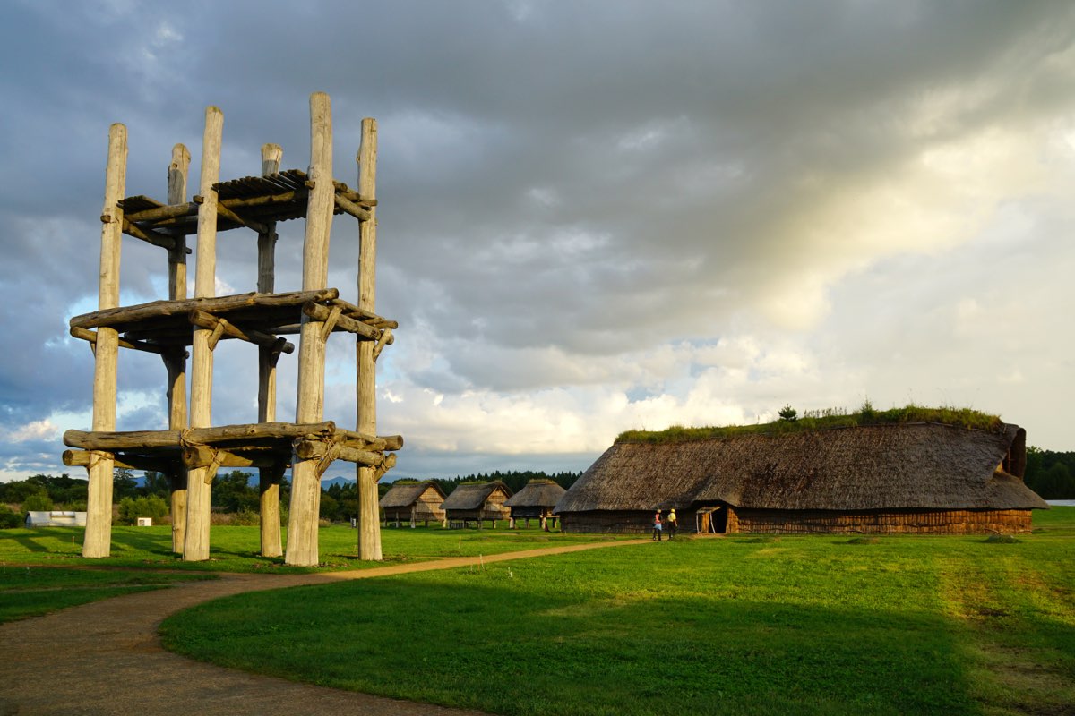 A tall open timber frame on six posts and a long thatched building on a grassy field at the Sannai-Maruyama site, with smaller huts in the distance