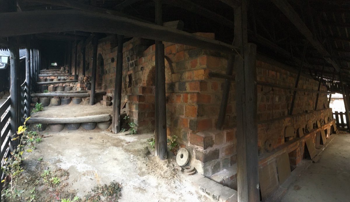 A long brick climbing kiln with arched openings, running up a slope under a dark wooden roof held up by wooden posts, with steps along its left side