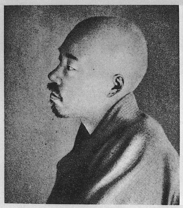 Black-and-white photograph of a man in profile facing left, with a shaved head and a moustache, wearing a dark kimono under a heavy padded outer garment, against a plain background