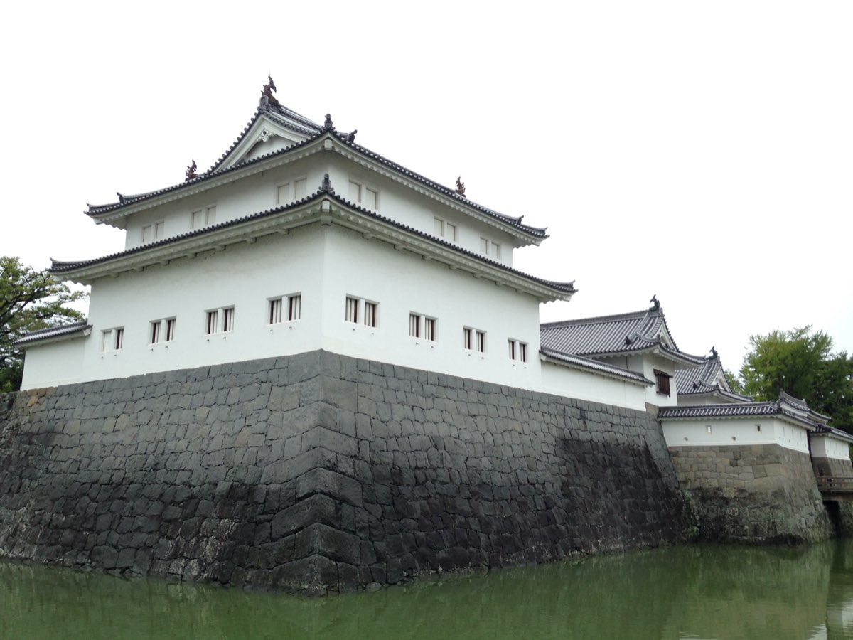 曇り空の下、緑色の堀から立ち上がる高く黒っぽい石垣の上に建つ、灰色の瓦屋根が2段に重なる白い隅櫓。右手には、低い石垣の上に瓦屋根の白い門の建物が続いている