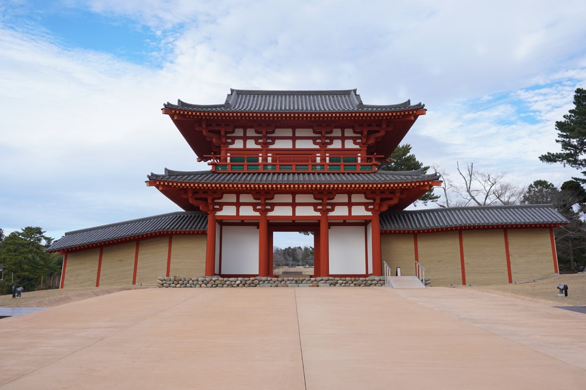 The reconstructed two-storey south gate of Tagajō, with red-painted pillars, white walls and grey tiled roofs, flanked by long roofed earthen walls, seen from a wide paved approach under a partly cloudy winter sky