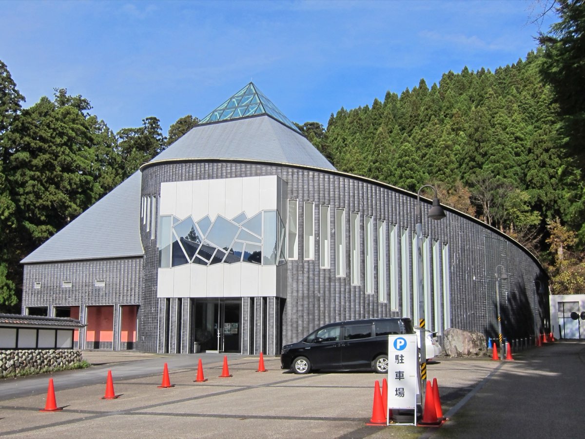 2018年に撮影された、立山博物館の建物。曲線を描く灰色の壁と、ガラスの天窓をのせた三角屋根が、木々の茂る斜面の前に建つ